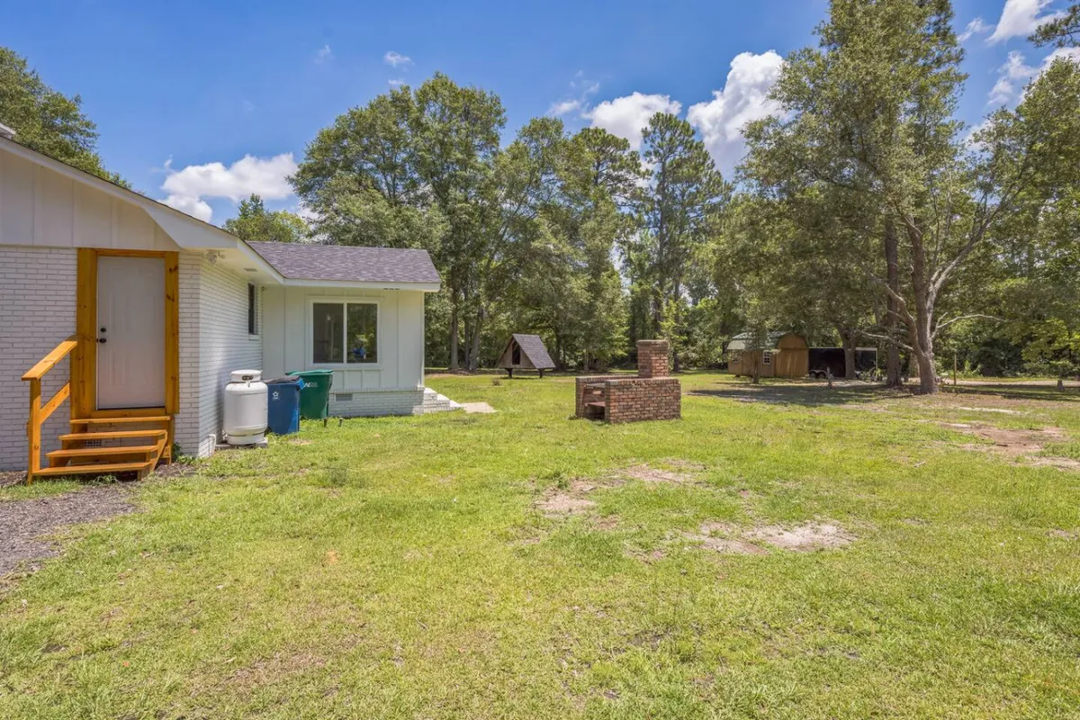 The image showcases a welcoming outdoor setting with wooden steps leading up to the house, a brick fire pit, and lush trees providing a peaceful, green backdrop. There's a sense of tranquility and opportunity for relaxation or productivity on the spacious lawn or shaded seating areas.
