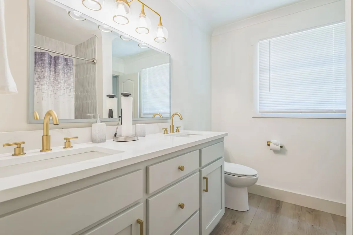 A spacious and bright bathroom featuring a double sink with elegant gold fixtures, a large mirror, and tasteful lighting. The setting suggests a clean, modern, and inviting space designed for self-care and relaxation.