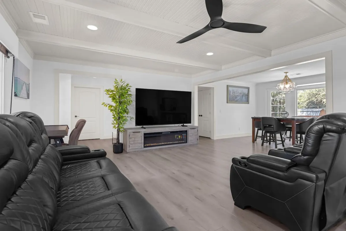 A spacious and modern living room with plush black leather sofas arranged for both relaxation and conversation, a large flat-screen TV perfect for movie nights, an indoor plant adding a touch of nature, and an adjacent dining area with a chandelier. Natural light filters in, making the space feel open and connected to the outdoors.