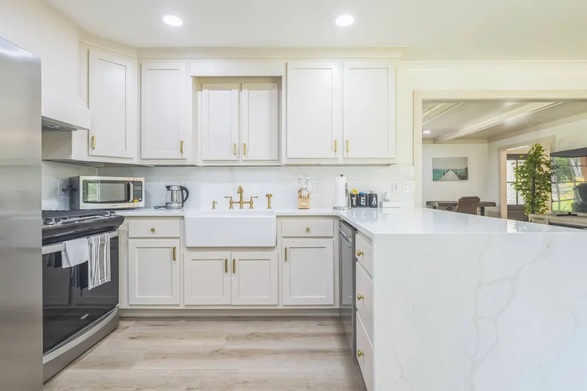 A kitchen scene showcasing a farmhouse sink with a gold faucet, marble countertops, stainless steel appliances, and light wood flooring. The overall design feels both elegant and homey, ready for everyday cooking or special celebrations.