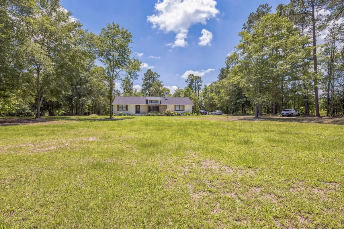 This exterior image showcases the house nestled among mature trees, with an open lawn that provides a sense of tranquility and space. The natural surroundings create a peaceful setting, ideal for rest and relaxation after a busy day. The ample outdoor area hints at practical benefits such as easy parking and space for families to gather or unwind.
