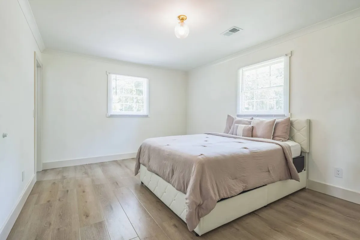 A tranquil bedroom scene showcasing a bed with soft bedding, warm wooden flooring, and generous natural light streaming in from large windows. The setup exudes a cozy, peaceful environment ideal for unwinding after a busy day.