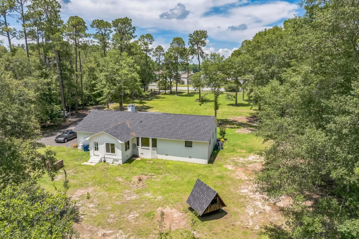 A spacious backyard shaded by mature trees, complete with a play structure and ample grassy area for children to play or families to gather. The back of the house and porch are visible, set against a peaceful natural backdrop.