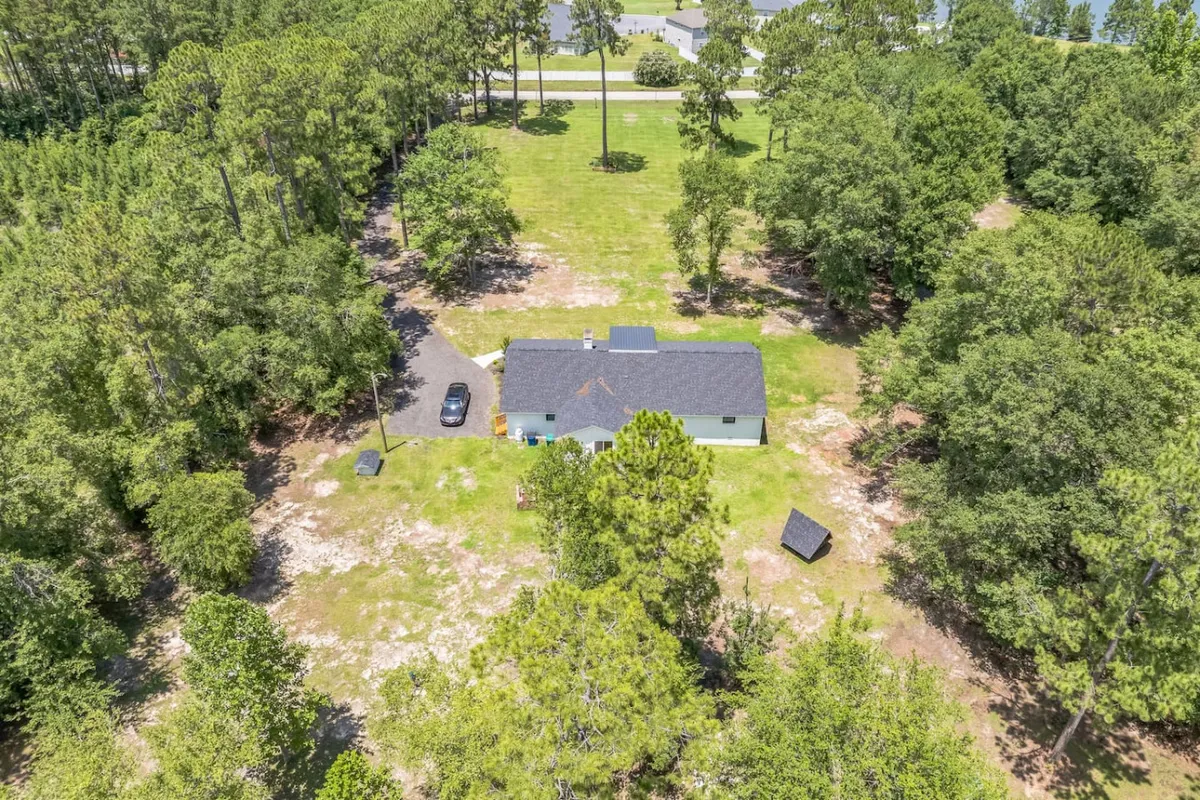 A wide-angle view of the house set among tall trees, with a prominent driveway providing ample parking. The home is framed by natural landscaping and open skies, underlining the sense of seclusion and space.