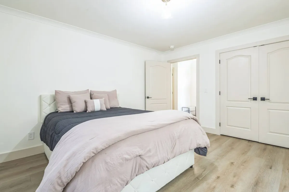 A serene bedroom with a plush bed dressed in decorative pillows, positioned to capture natural light spilling through double doors that lead to another room. The neutral color palette and uncluttered space underscore a restful and calming atmosphere.